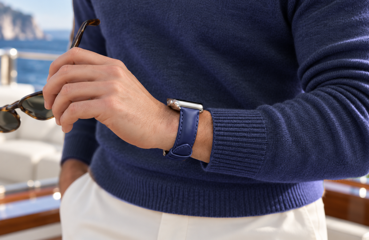 Man in a navy jumper holding sunglasses aboard a yacht, wearing a WsC Defiant blue leather Apple Watch strap with standard non-diamond silver hardware.