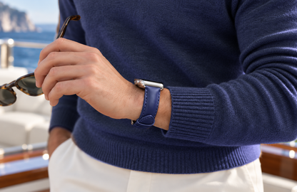 Man in a navy jumper holding sunglasses aboard a yacht, wearing a WsC Defiant blue leather Apple Watch strap with standard non-diamond silver hardware.