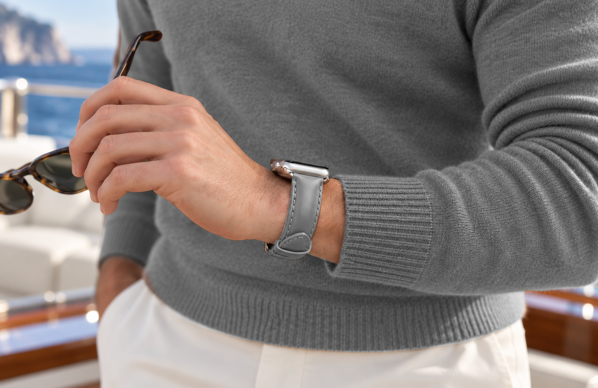Man in a grey jumper holding sunglasses and wearing a WsC Defiant grey leather Apple Watch strap aboard a yacht, with standard non-diamond silver hardware.