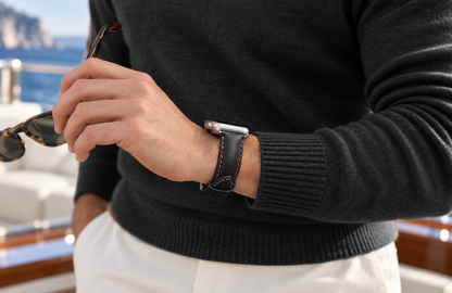 Man in a black jumper holding sunglasses aboard a yacht, wearing a padded WsC Defiant Pink Edition black leather Apple Watch strap with pink stitching and standard non-diamond silver hardware.
